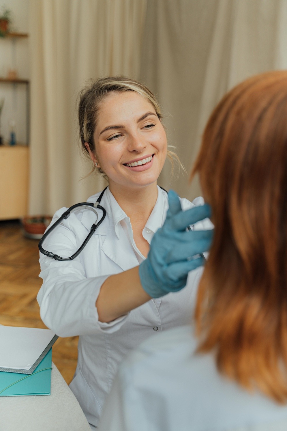 Female doctor smiling, wearing a stethoscope and blue rubber gloves analyzing closely a patients face. The patient's back is to the camera.