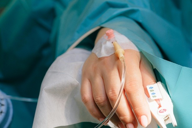 A close up image of a hospital patients hand with an IV in laying in a hospital bed wearing a teal patient gown.