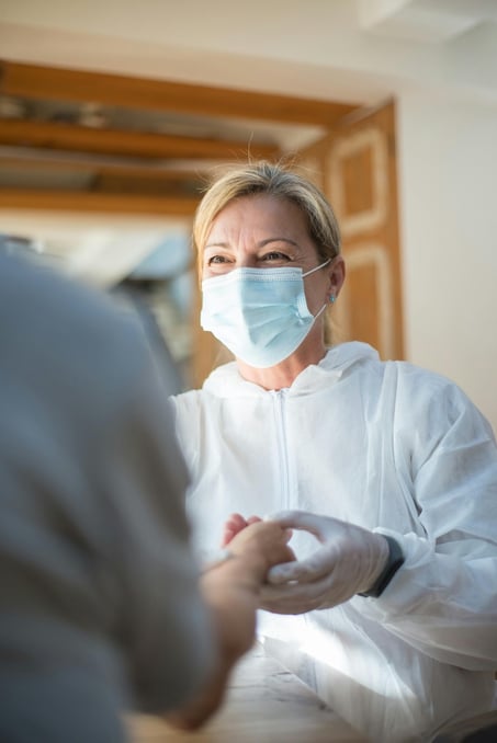 A woman smiling looking at her patient in a hospital. She is wearing a mask over her face, a white hoodie and white rubber gloves