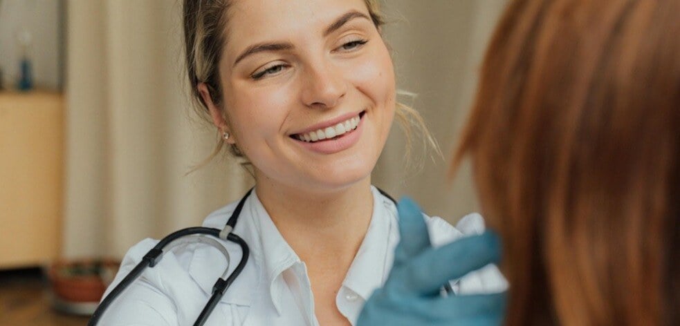 In image of a female healthcare worker looking after a patient. She is smiling and wearing a stethoscope around her neck. 